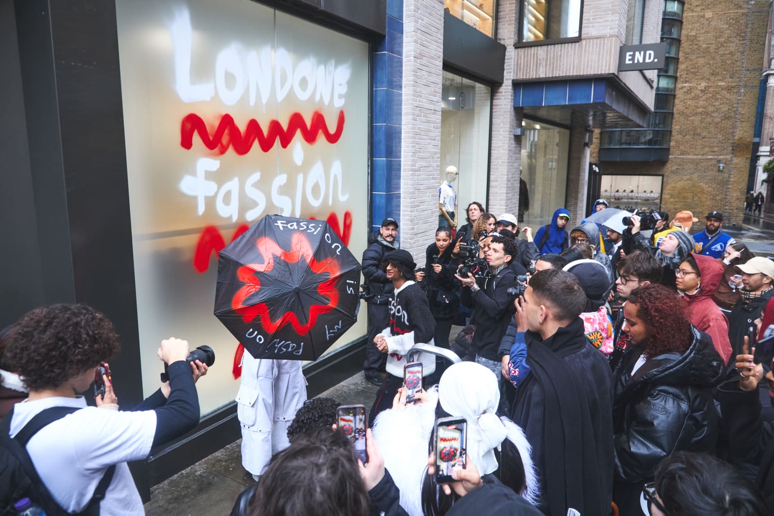 A crowd gathers outside a shop window that has been painted with white and red graffiti. 