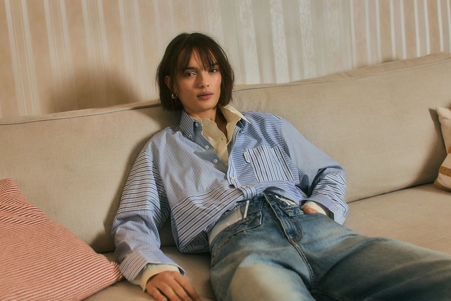 A woman with brown hair sits on a sofa wearing a blue and white striped shirt. 