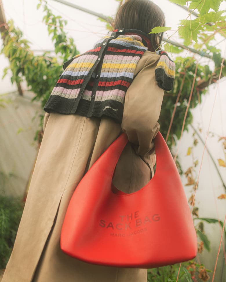 A woman faces away from the camera in a green house holding a red hand bag. 