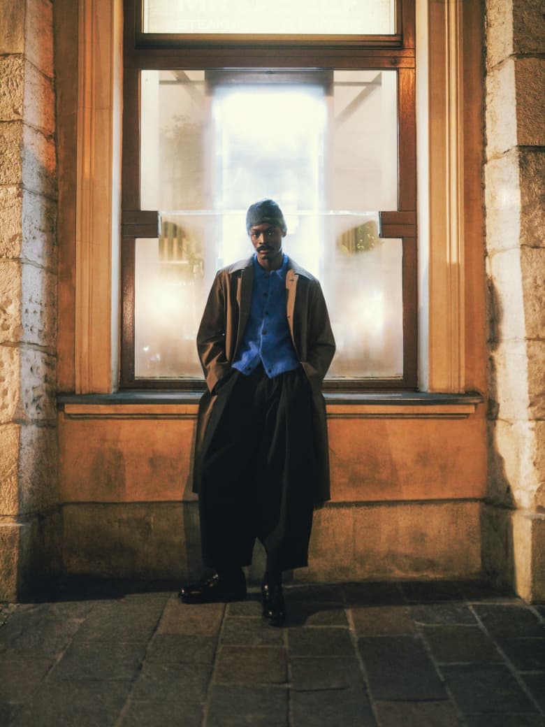 A man wearing Dries Van Norten coat and jumper, Y M C trousers and And Wander beanie.