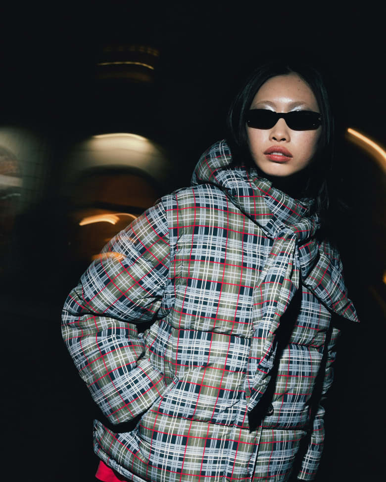 Woman stood wearing Miu Miu sunglasses, Acne Studios T-shirt and Damsen Madder puffer jacket.