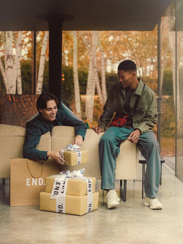 Two men unwrapping presents from End, wearing Salomon sneakers, C.P. Company jacket and Stone Island crossbody bag. 