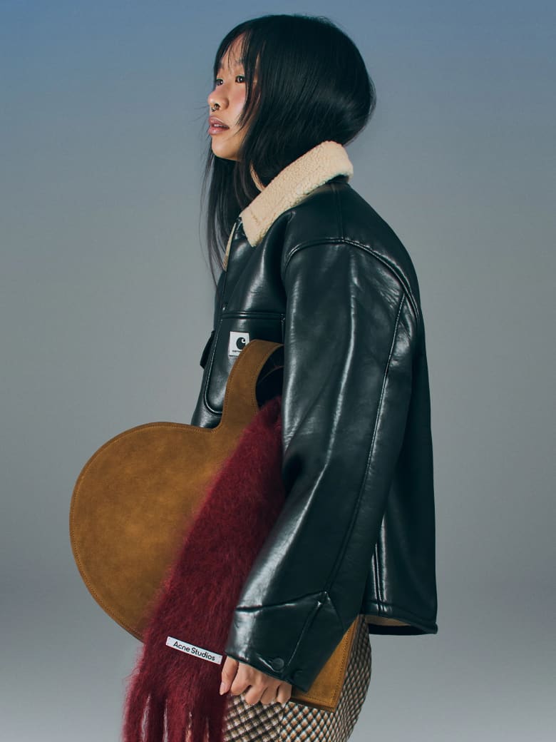 A woman wearing a black leather jacket and holding a brown heart-shaped clutch bag. 