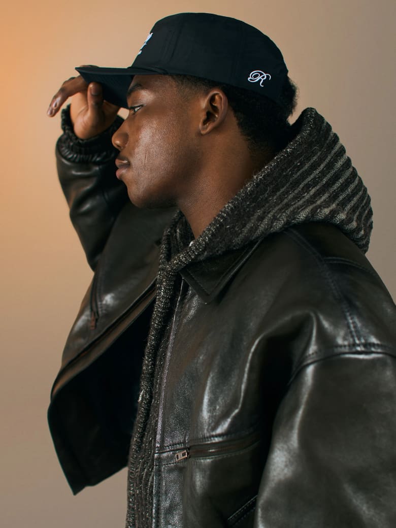 A man wearing a black leather jacket and a cap. 