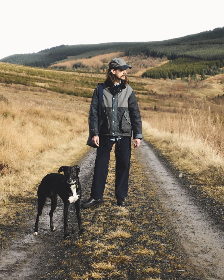 A man looking to the right standing on a public footpath with a greyhound on a lead. 