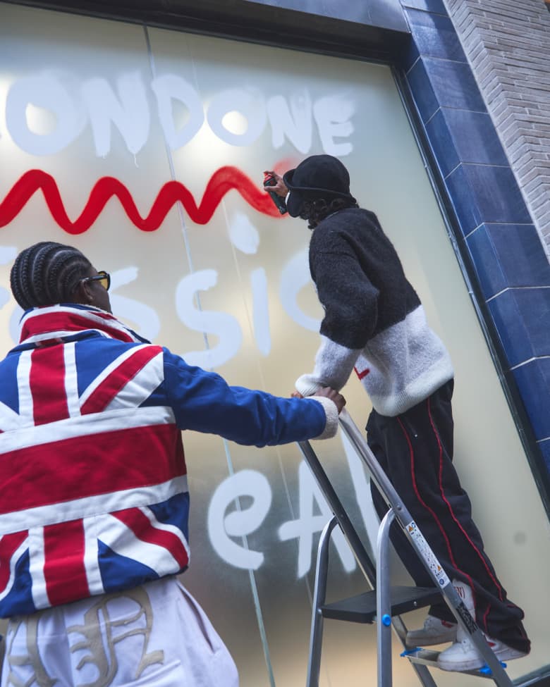 A man spray paints the word "London" in white paint on a shop window. 