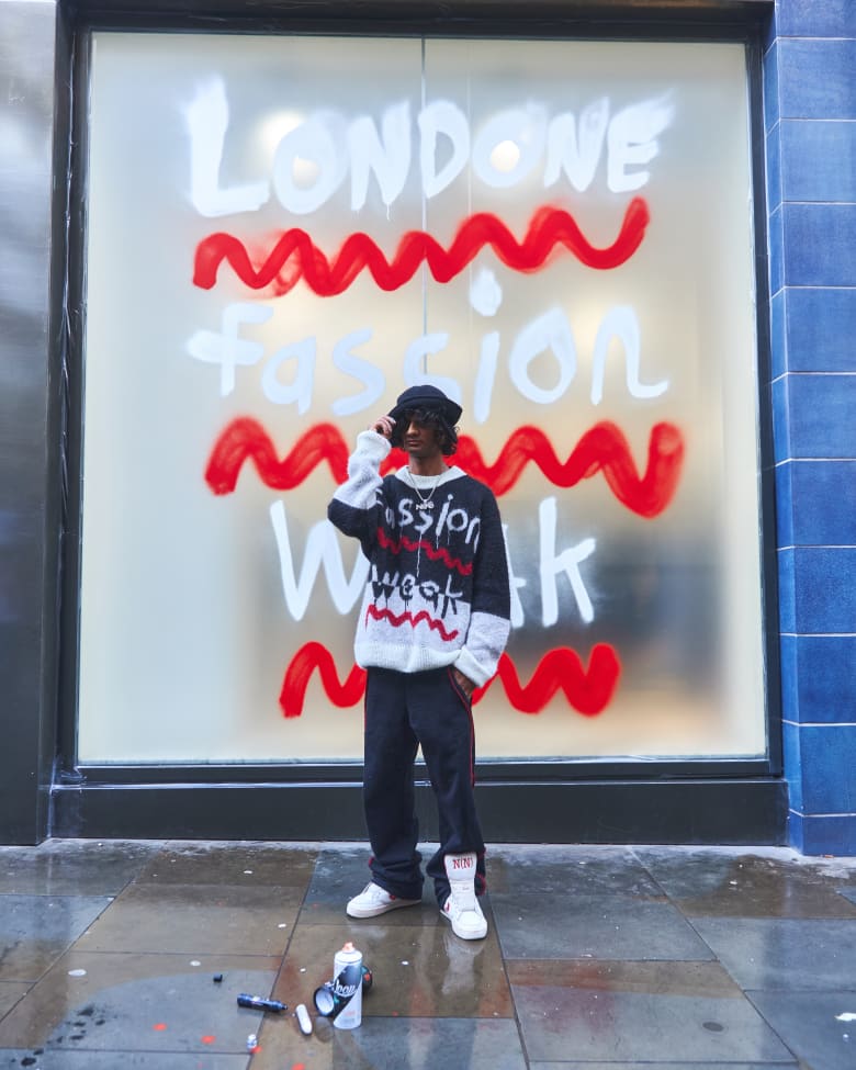 A man stands in front of a shop window that has been spray painted to say "Londone Fassion Weak".