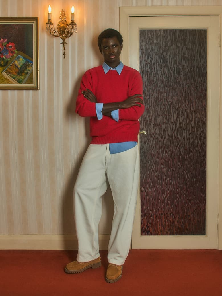 A man wearing a red knitted jumper stands next to a wall in a mid-century living room. 
