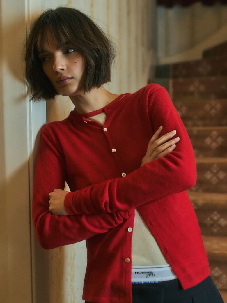 A woman wearing a red cardigan stands next to a wall in a mid-century living room in front of the stairs.
