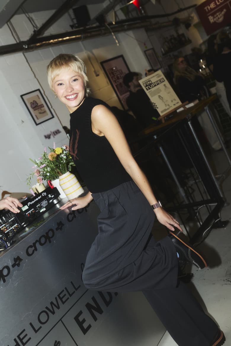 A woman with short blonde hair standing by DJ decks holding her shoe.
